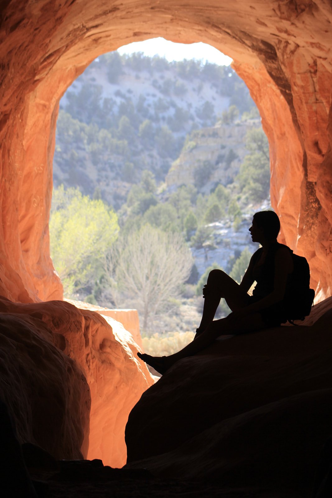 A silhouette of a woman sits in a tunnel made of red rocks overlooking a view.