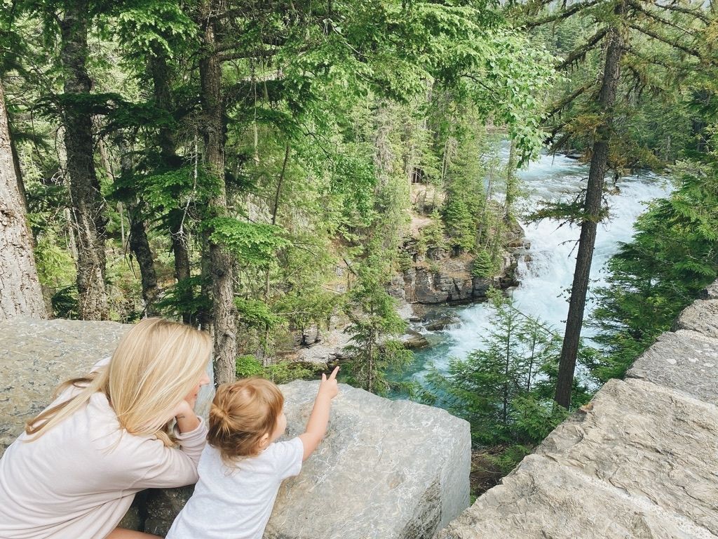 Blonde woman with toddler pointing at a river and forest in Glacier National Park.