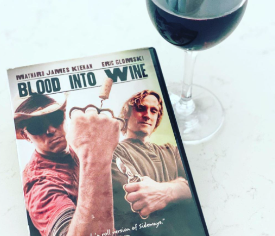 A copy of the documentary "Blood into Wine" with musician Maynard James Keenan on the cover and glass of wine from Caduceus Cellars sit side-by-side atop a marble table.