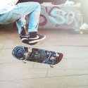 Person in blue jeans and a white shirt doing a trick on a colorful skateboard in front of a wall with graffiti