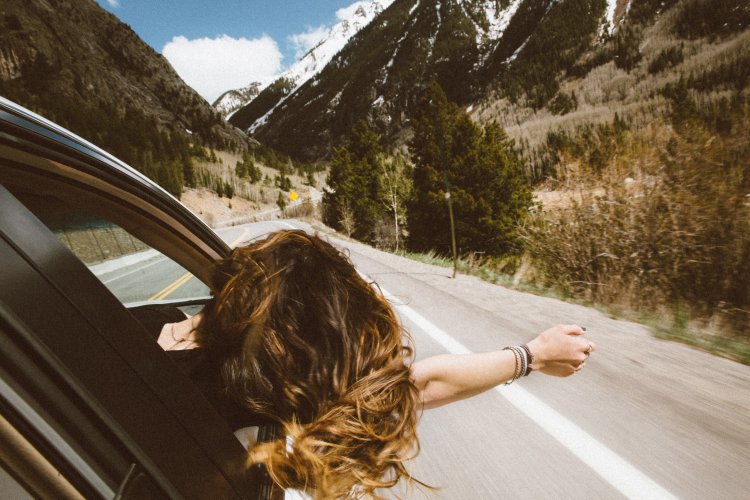 Woman stretching out the window of a car, letting her hair blow in the wind.  The vehicle is driving up a scenic mountain bi-way. 