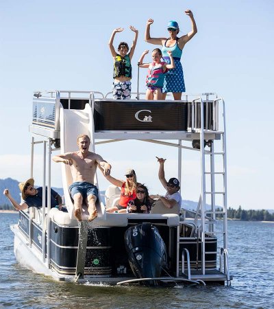 group of eight people riding a double decker pontoon boat on a lake and a man sliding down the boat’s water slide at Tamarack Resort