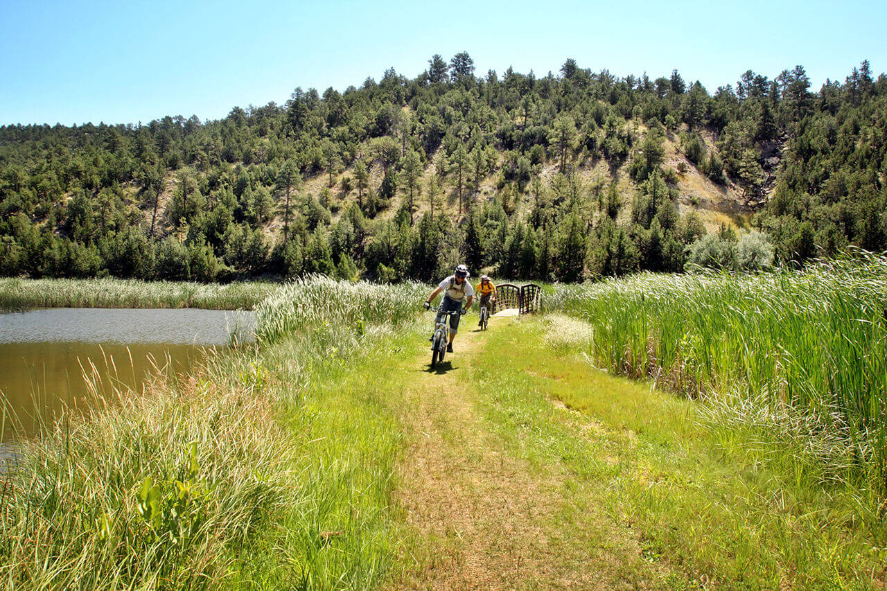 Two mountain bikers ride on a trail alongside water