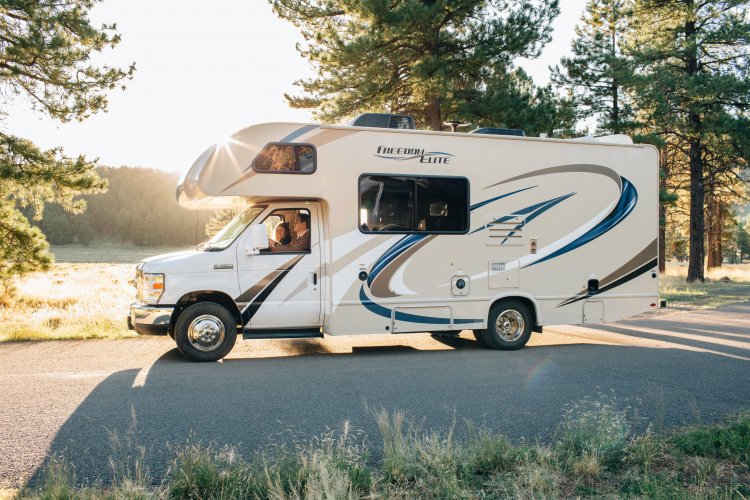 Couple driving an RV on a road flanked by tall trees.