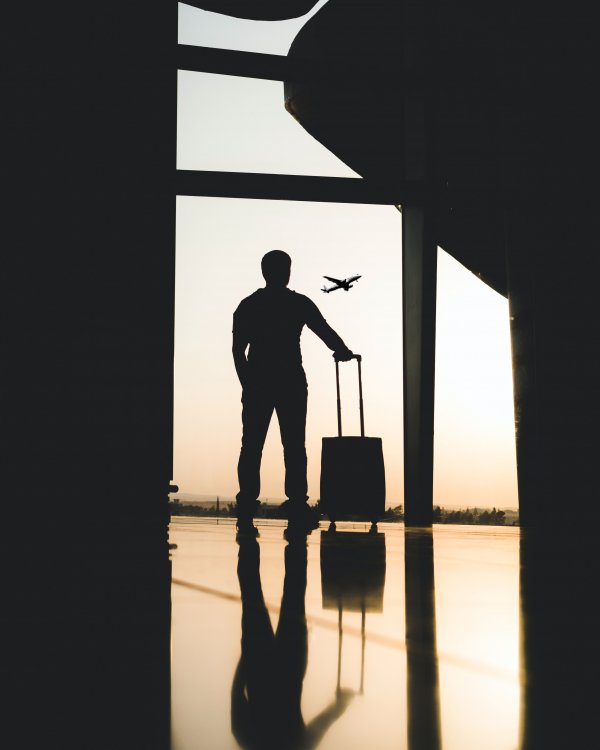 Man in silhouette with a suitcase looking out as a plane takes off in the distance